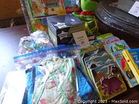 Photo of the lot showing bottle warmer box, baby toys including a green frog plush, board books, and zippered bag with baby clothes.