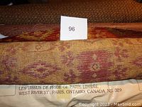 Close-up image of partial bolt of upholstery fabric showing woven multi-colored muted red and beige pattern. Also visible is label indicating fabric brand Les Tissus de Pride of Paris, Ontario, Canada.