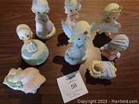 Seven Precious Moments Christmas figurines arranged on a wooden surface showing angels, children, and nativity themes. Includes a small music box and a lantern that needs repair.