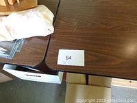 Two folding tables placed side by side showing the wood-grain laminate tops with a paper lot number 64 on one table
