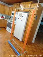 Full view of the clothing rack standing in a kitchen area, showing orange foam padding on the vertical pipes and blue foam on the horizontal base pipe.