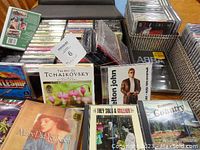 Photo showing a collection of CDs, cassettes, and DVDs. Visible album titles include 'True Tales of Prince Edward County', '3 Hits of the Swing Bands', Elton John CD, 'Tchaikovsky' compilation, 'Mary Did You Know?' book with music CD, and a Battleship computer game CD-ROM.