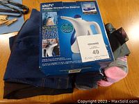 Photo of the Shift brand portable wrinkle free steamer in its original box with surrounding socks and pants visible under it.