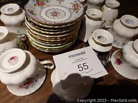 Top-down image of stacked Royal Albert saucers and cups around a MaxSold lot tag. Shows bottom stamps confirming Royal Albert brand and floral designs with gold trim.