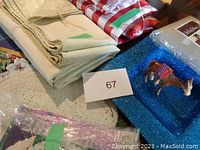 Photo showing several folded linens including beige or white tablecloth or placemats, red checkered fabric items, blue sparkly dish, and packaged white paper doilies.