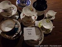 Overview of various tea cups and saucers arranged on a table, showing different floral patterns and gold trim, including dark blue Royal Stafford cup and saucer, and cream and sugar set in background