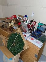 Wide view of lot showing various Christmas decorations including wreaths, ornaments, window décor, and light clips arranged on wood surfaces in basement.