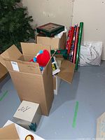 Boxes containing Christmas decorations including ornaments, bows, and wrapping paper rolls leaning against a wall.