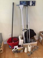 Photo showing mop bucket with wringer, white sponge mop, Swiffer cleaner, fly swatter, lint rollers, twine coils, hanging laundry accessory, and storage hooks.
