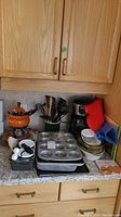 Overview photo showing coffeemaker, fondue pot with skewers, ceramic utensil jar, baking pans, glass dishes, oven mitts, and poultry holder on kitchen counter.