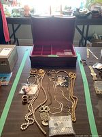 Open dark-colored jewelry box with velvet lining and compartments, placed behind assorted costume jewelry items including necklaces, rings, and bracelets.