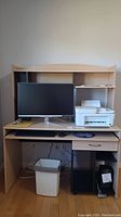 Front view of laminate wood desk with computer monitor, printer, and trash can placed around and on it, showing desktop and shelf layout.