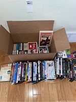 Box with multiple cassette tapes inside and some DVDs arranged on the floor, showing a variety of titles.