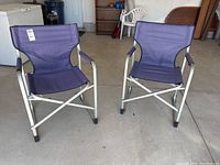 Full view of the two purple fabric folding chairs with silver metal frames and fabric arm covers, showing their structure and design.