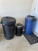 Three garbage bins side by side: blue Rubbermaid bin with lid and wheels, black DPI bin with lid and bungee cords, black metal bin with liner inside