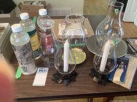 Photo of two glass oil lamp chimneys, two white candles with metal holders, and bottles of lamp oil on a wooden table.