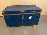 Front view of blue metal steamer trunk with lock and side handles on concrete floor.
