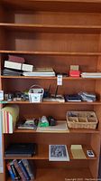 Photo showing the wooden shelving unit with multiple shelves holding stacks of paper, staplers, pens, calculators, baskets with small items, folders, and photo albums.