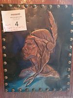 Close-up photo of copper plate portraying a Native American man in profile with a feather headdress, mounted on wood with metal rivets.