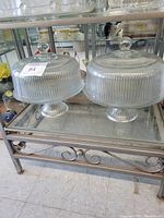 Two vintage glass dome cake plates with ribbed domes and pedestal bases on a metal and glass table.