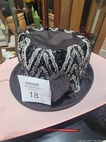 Full view of black women's hat with silver lined braided trim arranged in zigzag pattern around crown, sitting on table.