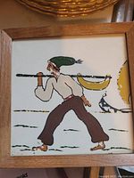 Front view of an Italian painted tile showing a man carrying a pole with a yellow object, framed on wood