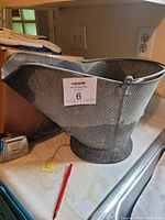 Angled side view of the metal ash bucket, showing the length, texture, handle, and pouring spout.