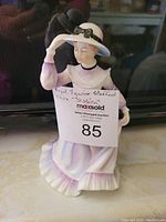 Front view of the porcelain figurine showing the lady in a white and lavender dress with a wide-brimmed hat and a choker necklace, holding a sign with the lot number and description.