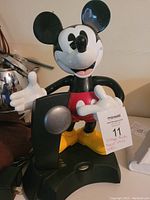 A color photo showing the Mickey Mouse statue base of the phone with open arms holding the handset cradle. The statue is in classic cartoon colors with red shorts and yellow shoes.