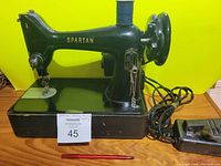 Side view of the black vintage Spartan sewing machine with electrical cord and foot pedal visible on wooden surface.