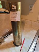 Full side view of the brass shell casing standing upright on a kitchen counter with a red pen for scale.