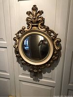 Full view of the ornate circular wood mirror hanging on a wall showing carved decorative frame with gold patina and pale green inner trim.
