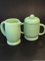 Front view of green jadeite glass creamer and lidded sugar bowl with handles, showing ribbed texture and pale green color.