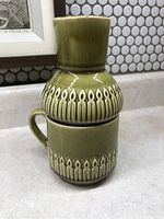 Green ceramic mid-century modern style mug stacked with matching decanter, both with glossy finish and raised pattern, no defects visible.