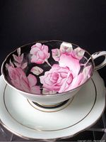 Close-up view of the teacup interior showing deep pink cabbage roses on a black background with mint green and gold accents on the edge.