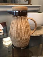 Full view of the vintage Corningware wicker coffee thermos, showing the body wrapped in tightly woven wicker, the chrome and wood lid, and wicker handle.