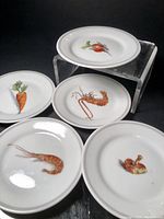 All five cocktail plates shown with distinct sea creatures and vegetable decorations on a black surface and a clear display stand.