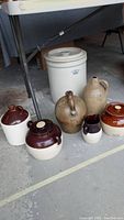 Full view of the lot items lined up on the floor under a table including the large crock, three jugs, two bean pots, and a pitcher.