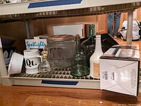 Full lower shelf view showing glass canister, green bottles, white ceramic carafe, crystal decanter, ramekin, mug, OGGI box and surrounding items
