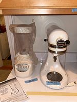 Photo showing Hamilton Beach blender on left with protective plastic cover and KitchenAid mixer on right on a white surface.