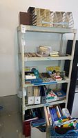 Full front view of five shelf plastic storage unit containing various office and paper items. Unit shows normal wear, scratches visible on plastic shelves and posts.