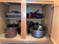 Full view of pots and pans inside kitchen cabinet showing various sizes and types including maroon enameled pot, stock pots, and pans