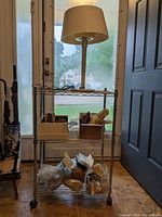 Metal rolling shelving unit with three wire shelves and caster wheels, holding several stones and rocks. Top shelf has white table lamp with drum shade.