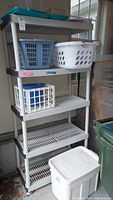 Full view of shelving unit with plastic containers including Rubbermaid tub with green lid on top, two laundry baskets on second shelf, milk crate and shoe boxes on third shelf, waste basket in foreground.