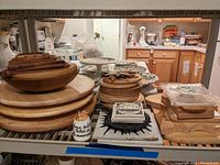 Wide view showing stacks of different sized and shaped wooden trivets, a wooden bowl, some ceramic coasters, and a small jar on a shelf in kitchen.