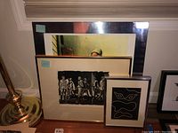 Three framed prints displayed leaning against a wall near a brass lamp base. Prints include a partially visible image of a person behind a window, a monochrome print of a group of figures, and an abstract line art print in black and white.