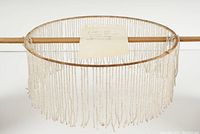 Front view of the glass bead fringe mounted on a circular frame, showing the full fringe and its hanging bead strands.