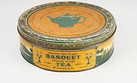 Top view of large round antique metal tea tin with green teapot design on the lid and decorative border. Yellow background with wear and some rust.