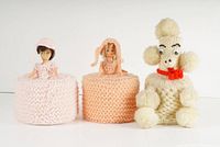 Front view of 3 crochet figural toilet paper roll covers: two with female dolls in pink and orange dresses, one poodle dog figure.