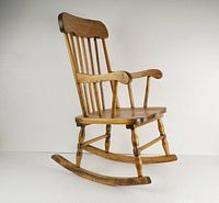 Front-left angle view of the small vintage wooden rocking chair showing curved arms, rockers and vertical spindle backrest.
